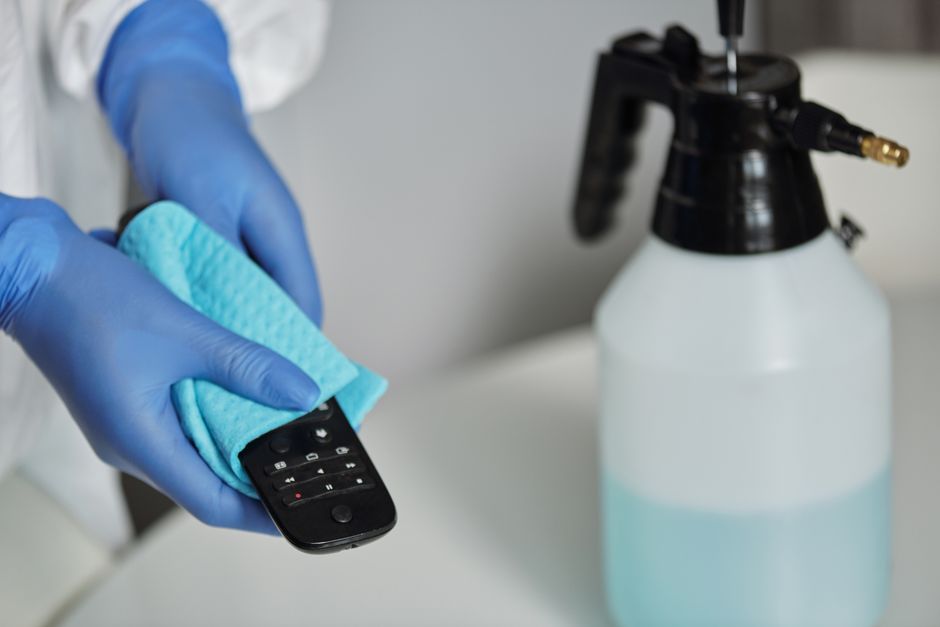 A close-up view of a hand wearing a bright pink rubber glove, holding a white spray bottle with a black nozzle, aimed at the edge of a pinkish-beige bathtub. Behind, there is a tiled wall with beige c