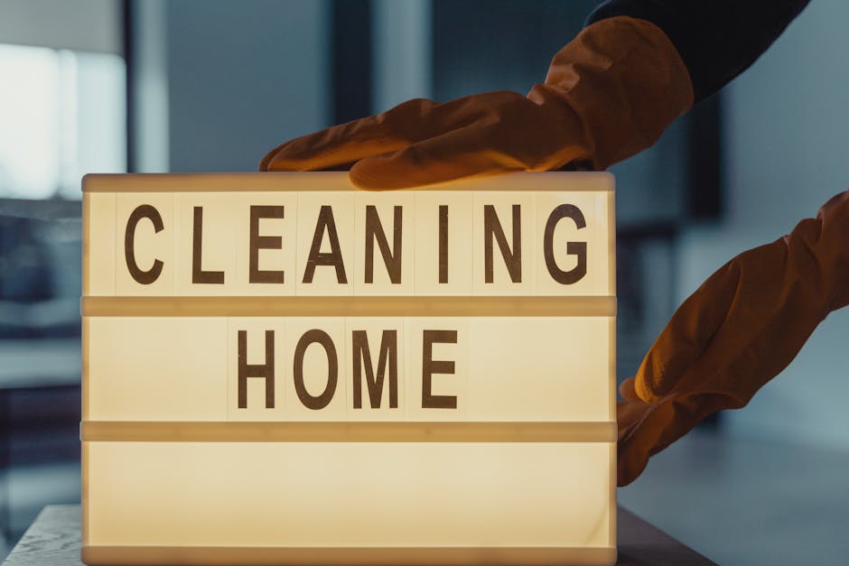 A person wearing orange cleaning gloves is adjusting a rectangular illuminated sign with black letters spelling 'CLEANING HOME' on a white background. The sign is placed on a flat surface in a well-li