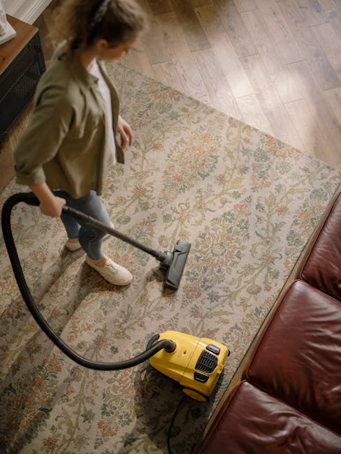 A woman with brown hair tied back, dressed in a light blue striped shirt and jeans, is kneeling on a patterned rug in a living room. She is using a blue cloth to clean the wooden floor near a dark blu